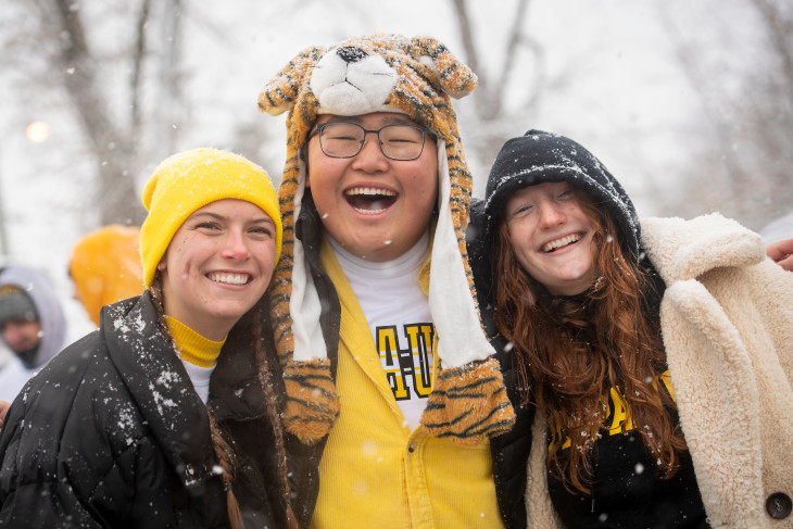 Three students smiling in the snow, one wearing a tiger hat