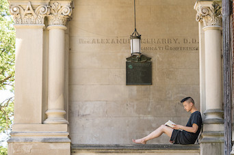 A student at DePauw sits along the steps of East College at DePauw University reading from a book. 